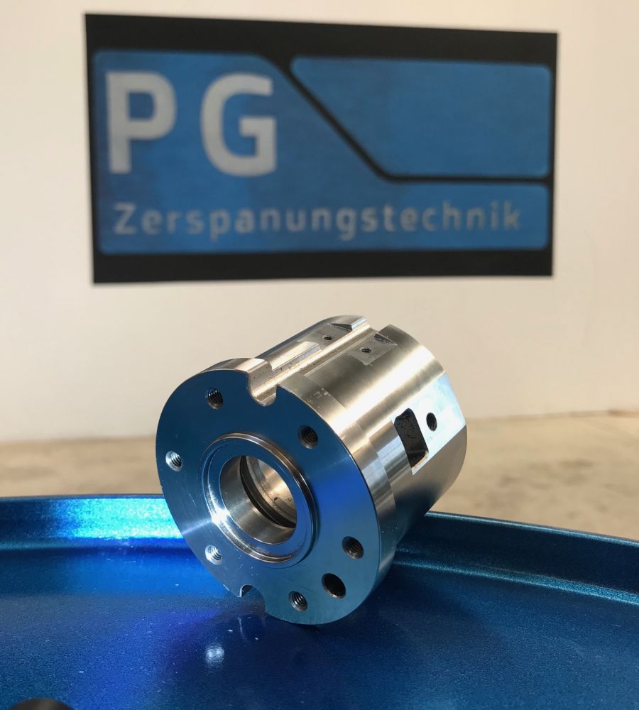 Metallisches Bauteil auf blauem Tisch vor PG Zerspanungstechnik-Logo im Hintergrund